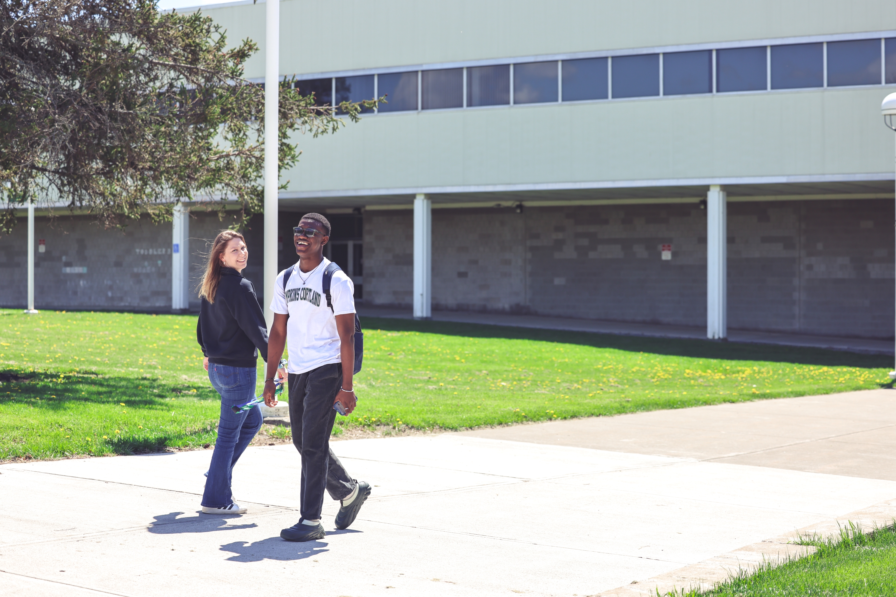 Two people walking around campus, laughing and smiling. There is green grass in the background and a tree. 